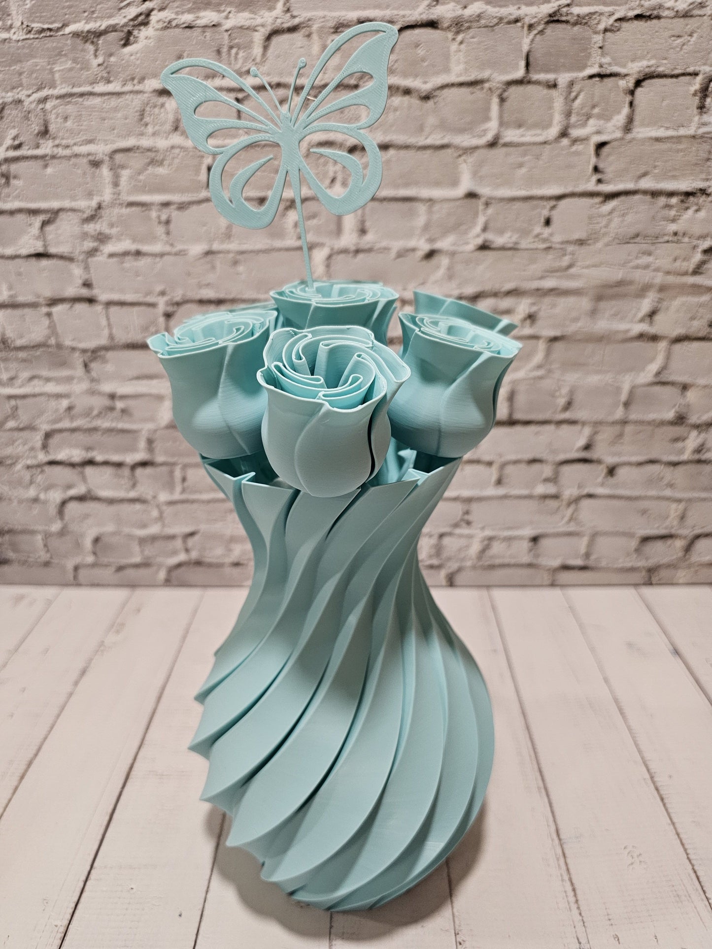 A light blue decorative bouquet sculpture made of layered paper-like roses in a twisted vase shape, topped with a butterfly cutout, set against a white brick wall and light wood floor.